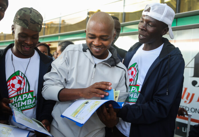 Football Kenya Federation President Nicholas Mwendwa (Middle) vibrantly engage some of the Kapenguria Heroes players, skimming through their travelling documents ahead of their check-in for their travel to London at the Jomo Kenyatta International Airport. - Bizna