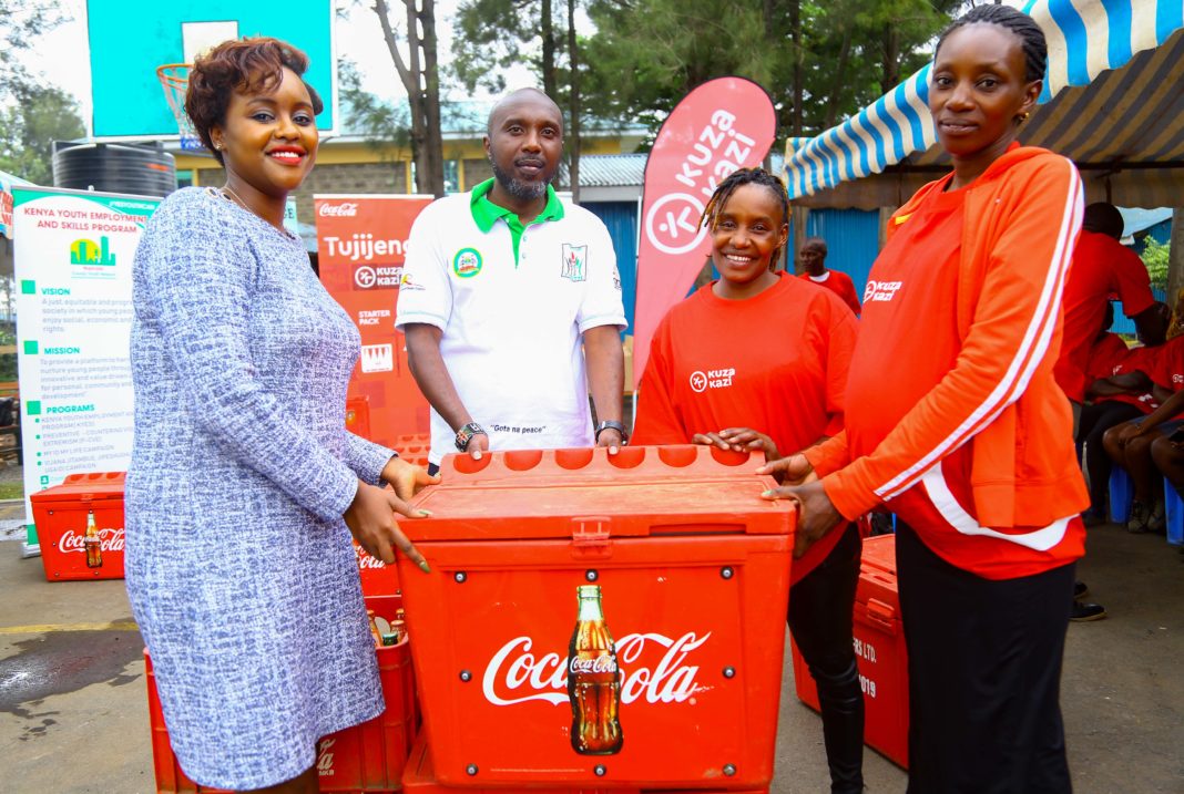 Coca-Cola Sustainability Coordinator Public Affairs and Communications, Victoria Macharia (Left), in company of One-Stop Youth Program Coordinator, Wainaina Muiruri (second left), present a Coca-Cola sales starter pack inclusive of a cooler box and crates of Coca-Cola beverages to Kuza Kazi beneficiaries, Wincete Karemi (Right) and Doreen Omwenga. - Bizna Kenya