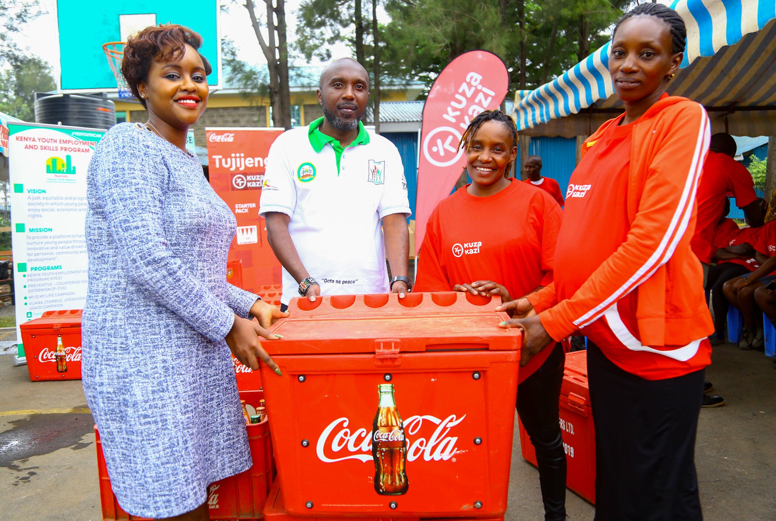 Coca-Cola Kuza Kazi Initiative Kits Youth In Nairobi County 1 Coca-Cola Sustainability Coordinator Public Affairs and Communications, Victoria Macharia (Left), in company of One-Stop Youth Program Coordinator, Wainaina Muiruri (second left), present a Coca-Cola sales starter pack inclusive of a cooler box and crates of Coca-Cola beverages to Kuza Kazi beneficiaries, Wincete Karemi (Right) and Doreen Omwenga. - Bizna Kenya